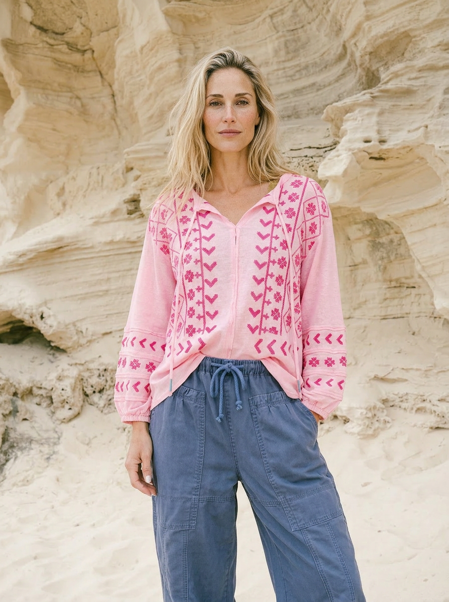 Woman wearing a pink embroidered top and blue pants standing against a rock wall.