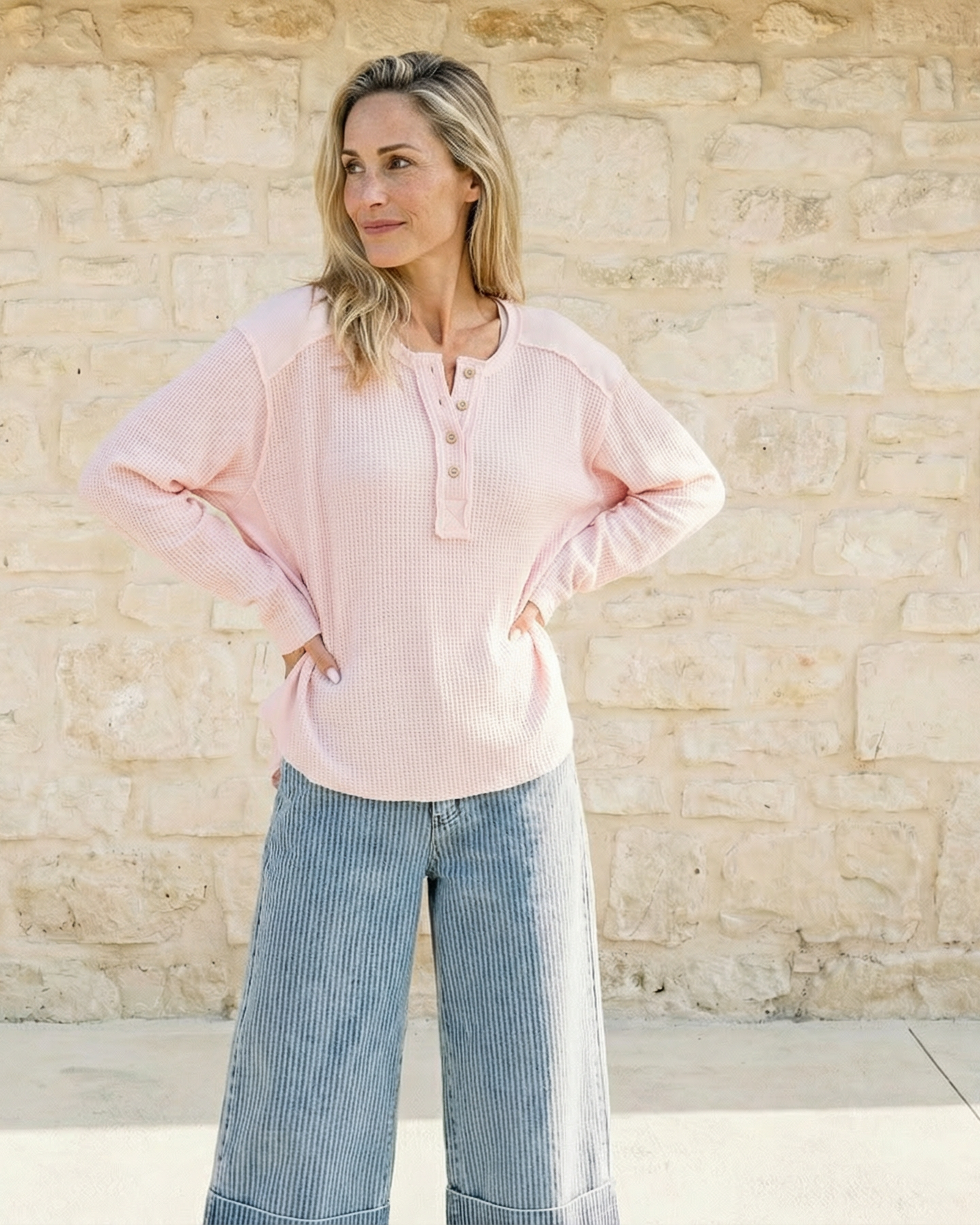 Woman wearing a light pink blouse and blue jeans standing against a stone wall.