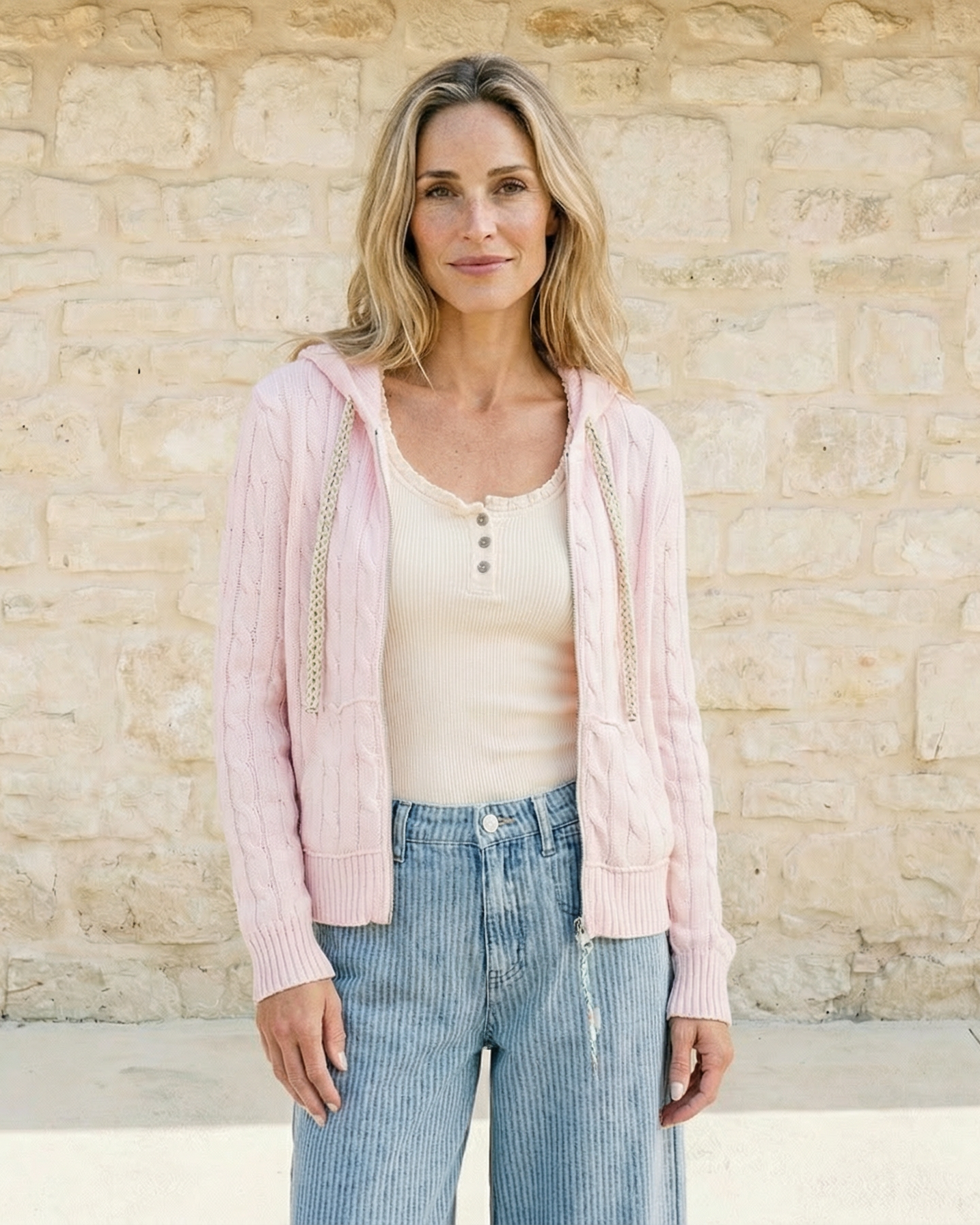 Woman wearing a light pink cardigan over a white top and blue jeans, standing against a beige stone wall.